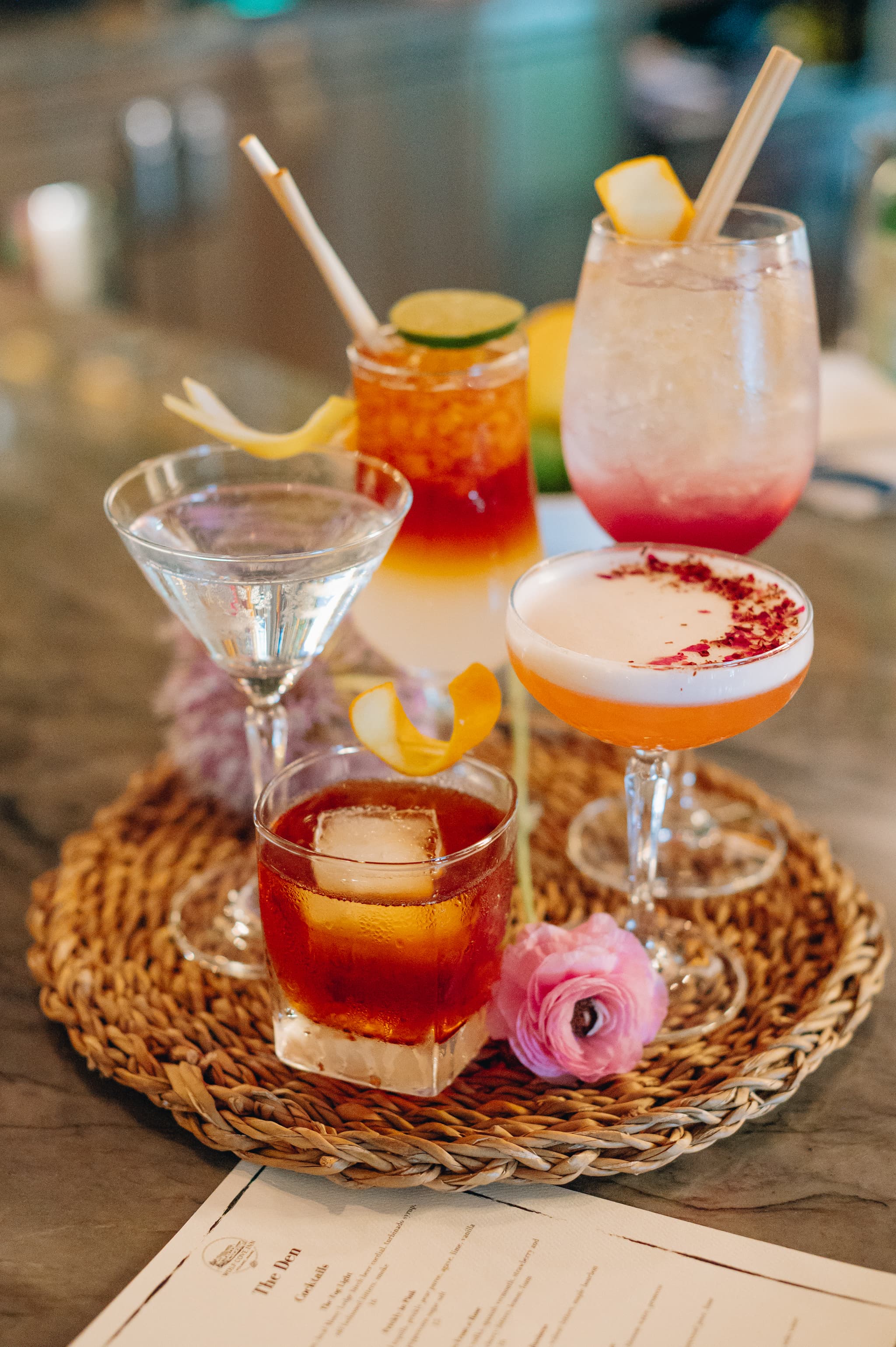 A selection of colorful cocktails arranged on a woven tray with a flower and a menu.