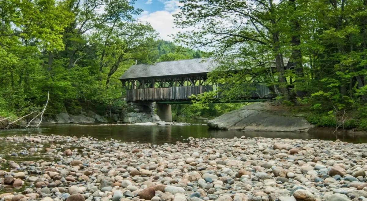 sunday river covered bridge in maine. rocky shore of a river with trees.