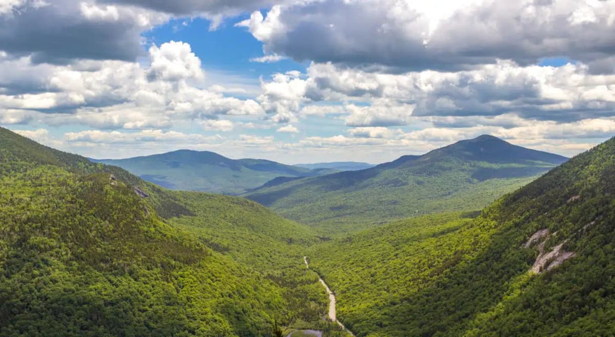 Distant view of Grafton Notch State Park with blue skies, white puffy clouds, green tree covered hills, and mountain peaks.