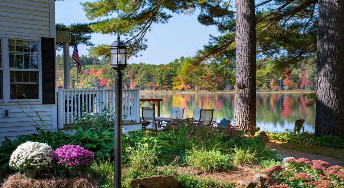 Side yard of white sided inn filled with colorful mums and plants, and a view of fall foliage reflecting in the lake in the background