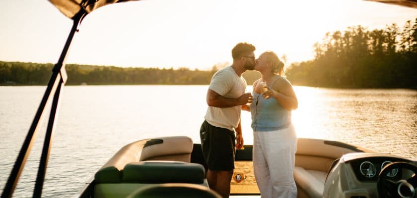 A couple sharing a kiss on a pontoon boat at sunset, with drinks in hand, calm lake water, and golden light behind them.