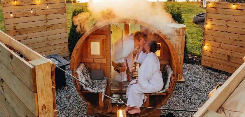 A couple sharing a romantic moment in a steaming outdoor barrel sauna with warm lights and cozy robes.