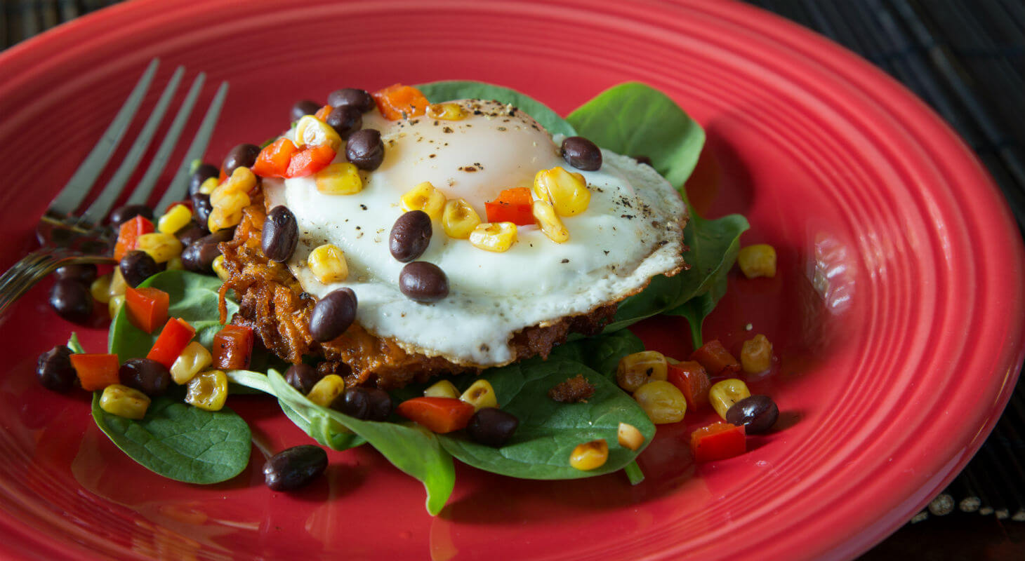 Eggs topped with corn and black beans over sweet potato hash brown and spinach on a red plate