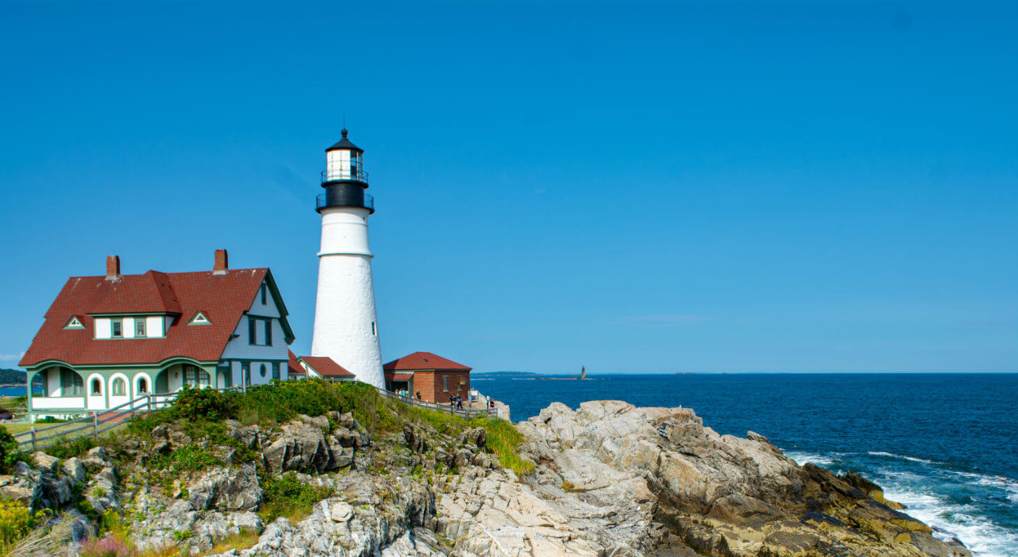 Lighthouse off the coast of Maine, above a beach with large rocks and next to a red-roofed two-story building