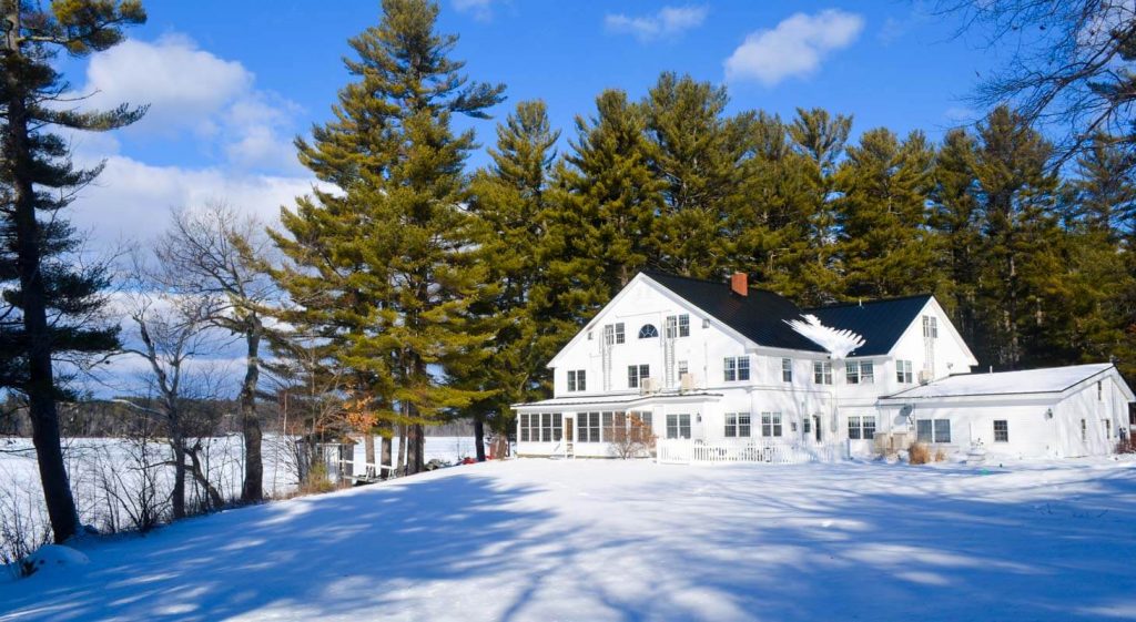 Exterior view of Wolf Cove Inn, white two story home surrounded by tall pine trees, blue skies and white snow