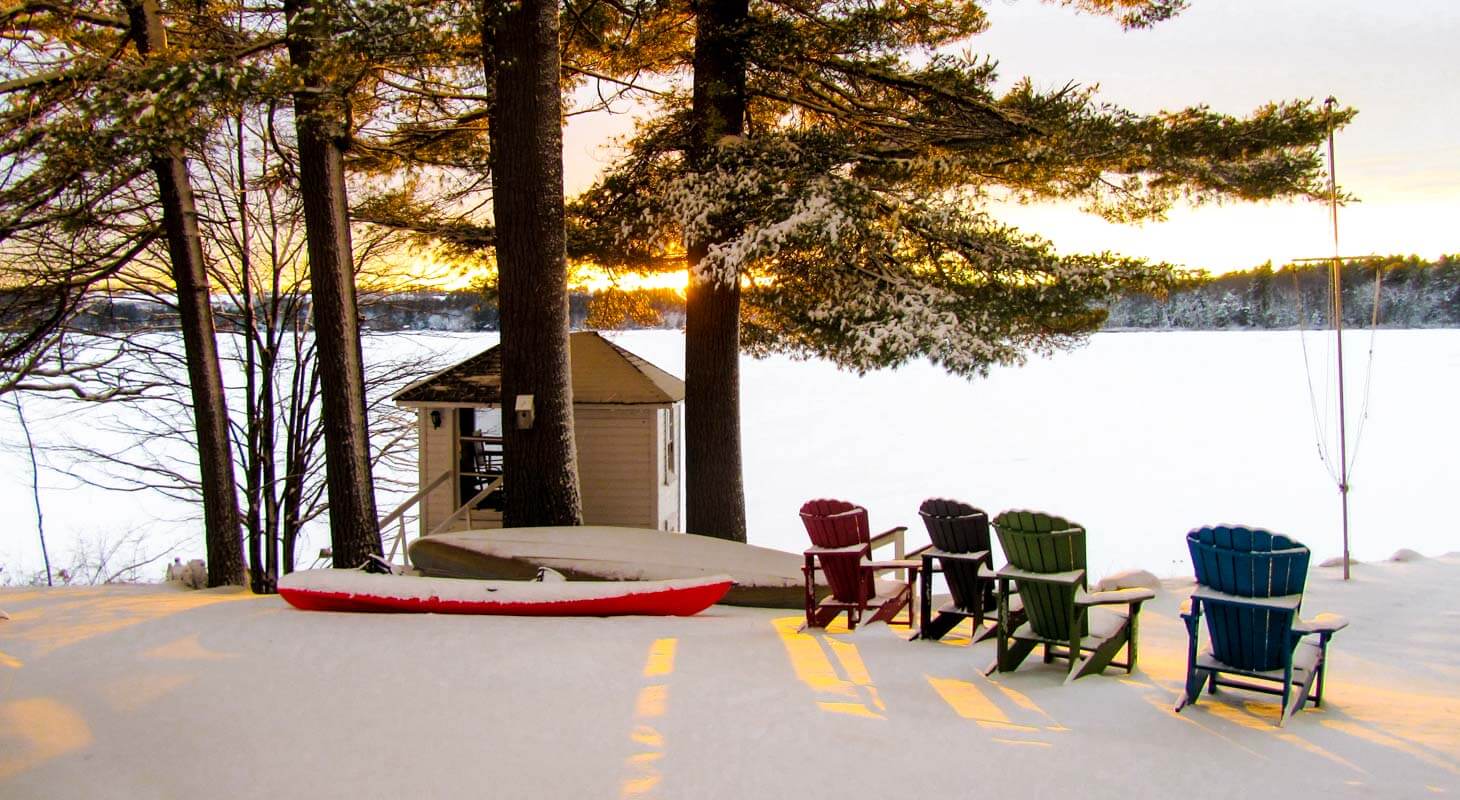Kayak and Adirondack chairs near a snowy, frozen lake at sunset.