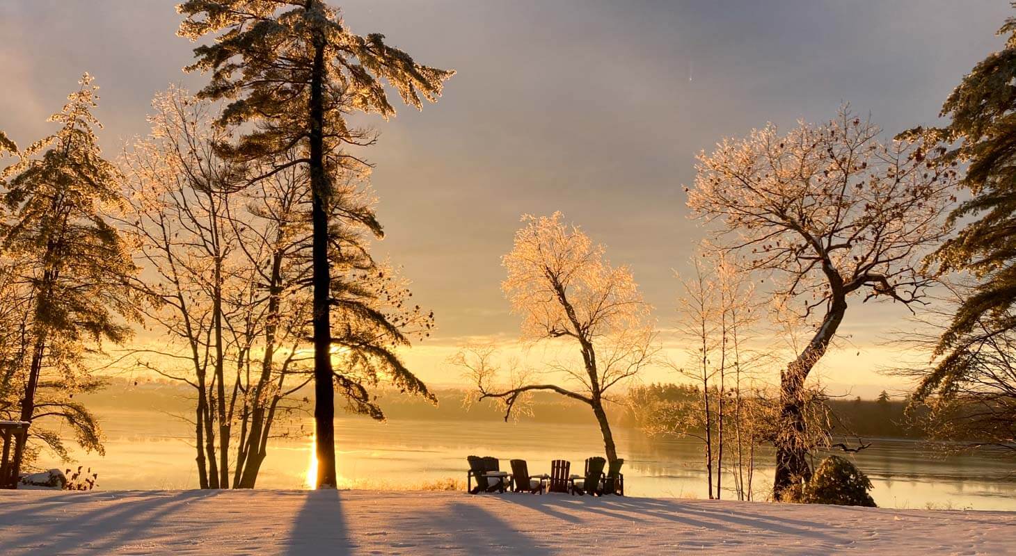 Sunset, trees and snow over frozen lake with chairs by a fire pit