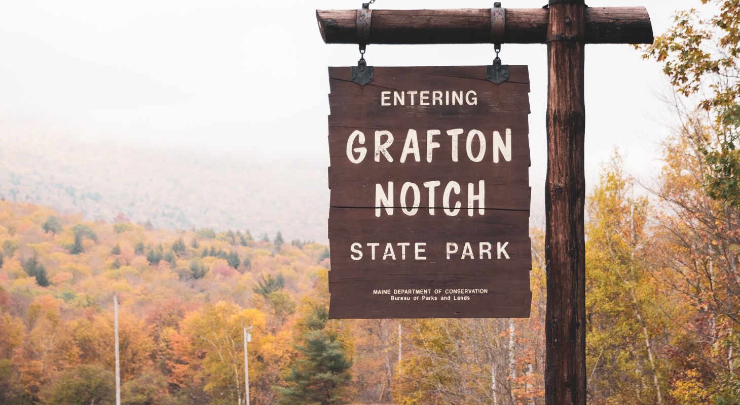 Wooden sign that says welcome to Grafton Notch State Park with fall foliage in the background