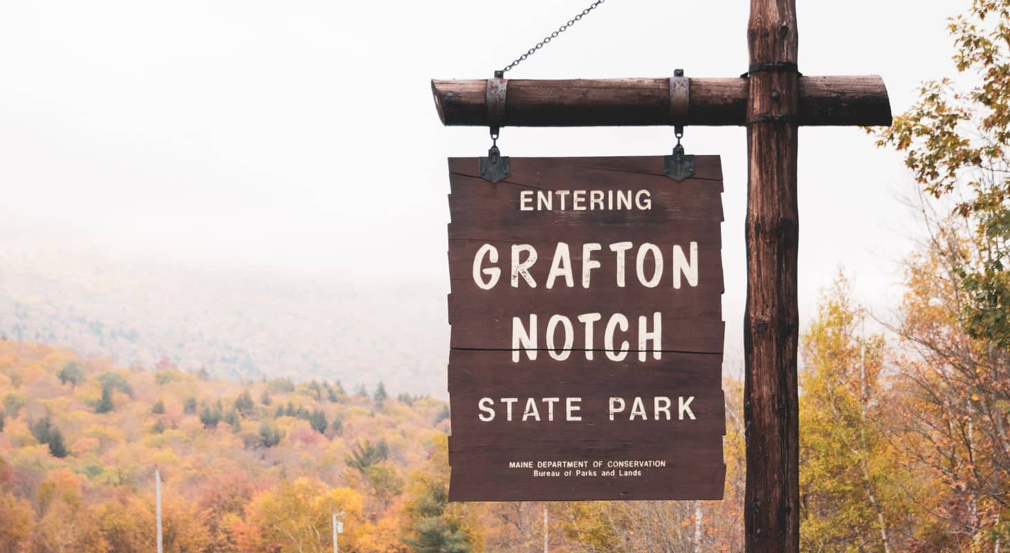 Brown wooden sign that says Entering Grafton Notch State Park with fall foliage in the distance