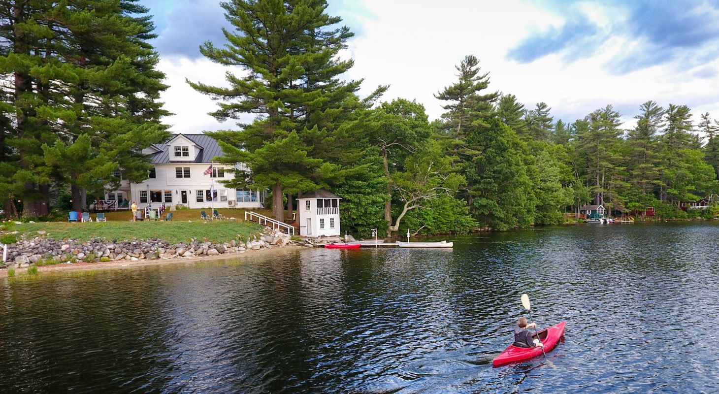 Person kayaking on Tripp Lake on a summer day, near the grassy shore of Wolf Cove Inn, a two story white bed and breakfast.